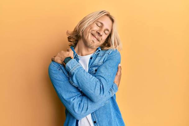 Homme caucasien aux cheveux longs blonds portant une veste décontractée en denim se serrant dans ses bras heureux et positifs, souriant confiant. amour de soi et soins de soi  - Photo, image