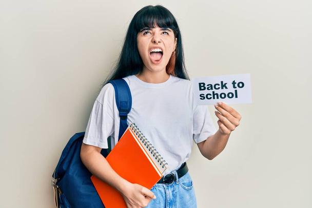 Jeune fille hispanique portant un sac à dos retenant la bannière de l'école en colère et folle hurlant frustrée et furieuse, criant de colère levant les yeux.  - Photo, image