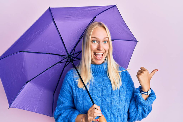 junges blondes Mädchen mit lila Regenschirm, Daumen zur Seite gerichtet, glücklich lächelnd mit geöffnetem Mund  - Foto, Bild