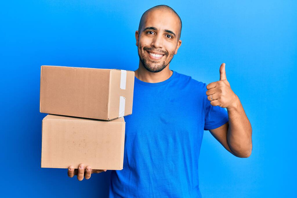 Hombre adulto hispano sosteniendo paquetes sobre fondo azul sonriendo feliz y positivo, pulgar hacia arriba haciendo excelente y signo de aprobación  - Foto, Imagen