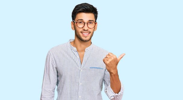 Young handsome man wearing casual clothes and glasses smiling with happy face looking and pointing to the side with thumb up.  - Photo, Image