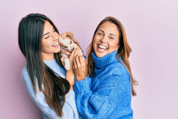 Beautiful hispanic mother and daughter smiling happy hugging little dog over isolated pink background. - Foto, Imagem