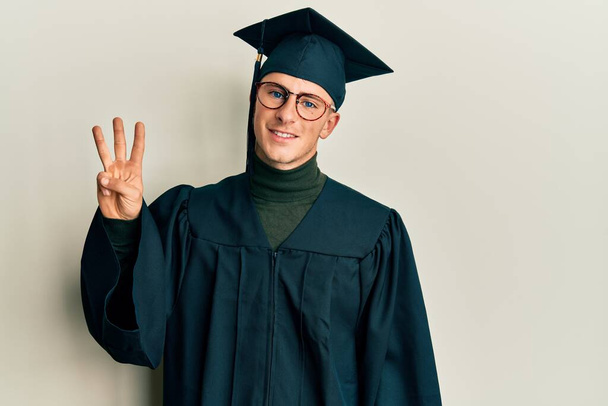 Jeune homme caucasien portant casquette de graduation et robe de cérémonie montrant et pointant vers le haut avec les doigts numéro trois tout en souriant confiant et heureux.  - Photo, image