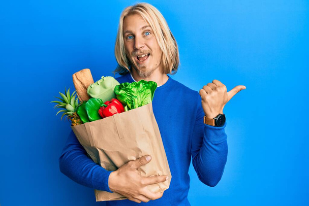 Белый молодой человек с длинными волосами держит бумажный пакет с продуктами указывая большой палец вверх в сторону улыбается счастливо с открытым ртом  - Фото, изображение