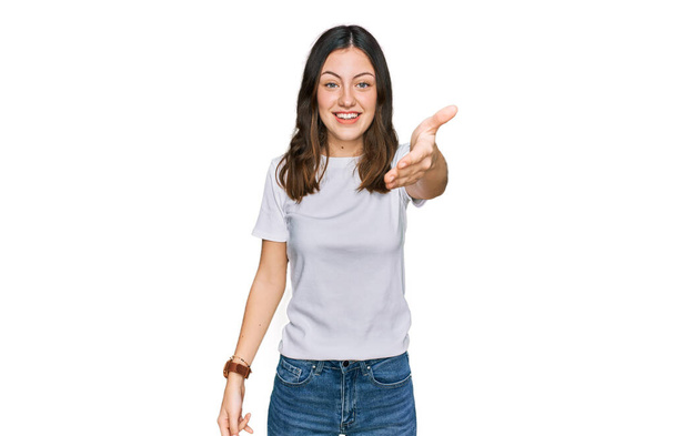 Mujer hermosa joven vistiendo camiseta blanca casual sonriendo amistoso ofreciendo apretón de manos como saludo y bienvenida. negocios exitosos.  - Foto, imagen