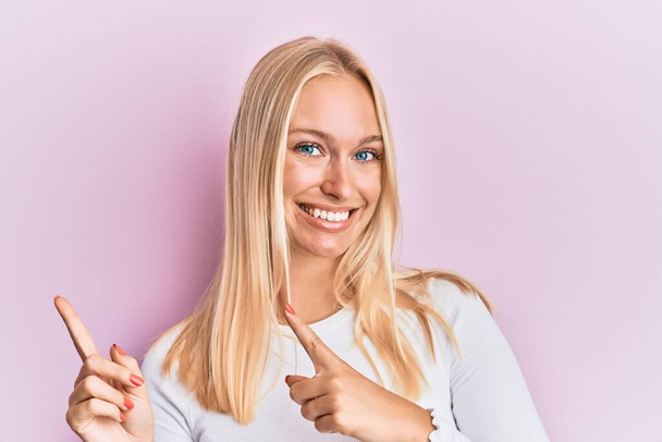 Mladá blondýna dívka na sobě ležérní oblečení s úsměvem a při pohledu na kameru ukazuje dvěma rukama a prsty na stranu.  - Fotografie, Obrázek