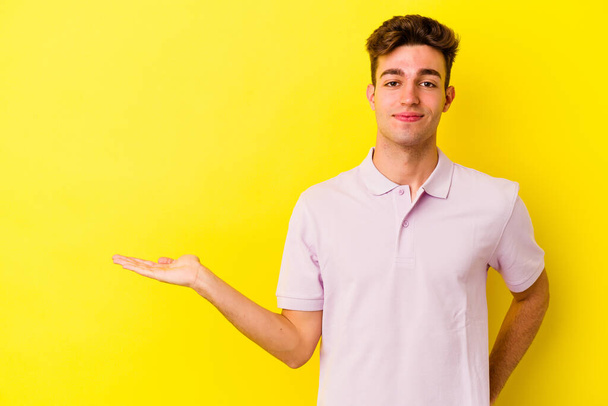 Молодий білий чоловік ізольований на жовтому тлі, показуючи простір для копіювання на долоні і тримаючи іншу руку на талії
. - Фото, зображення