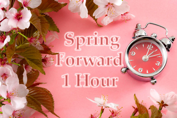 Uhren eine Stunde vor, Stern der Sommerzeit und Erinnerung an den Frühling vorwärts Konzept mit Uhr auf rosa Hintergrund mit Frühlingsblumen und Text - Frühling vorwärts 1 Stunde - Foto, Bild
