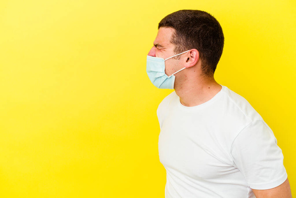 Junger kaukasischer Mann trägt einen Coronavirus-Schutz isoliert auf gelbem Hintergrund und schreit in Richtung Kopierraum - Foto, Bild