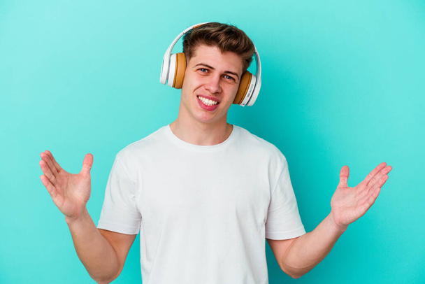 Giovane uomo caucasico che ascolta musica con le cuffie isolate su sfondo blu che riceve una piacevole sorpresa, eccitato e alzando le mani. - Foto, immagini