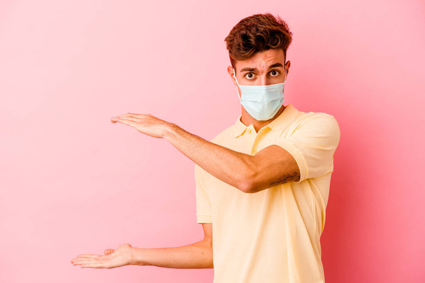 Jovem caucasiano vestindo uma proteção para coronavírus isolado em fundo rosa chocado e espantado segurando um espaço de cópia entre as mãos. - Foto, Imagem