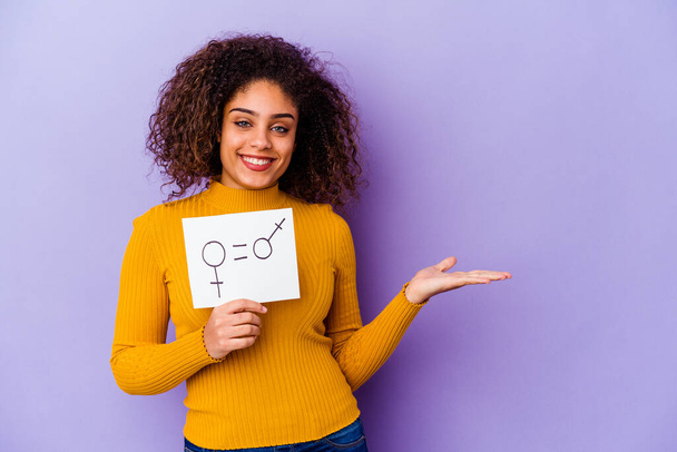 Joven mujer afroamericana sosteniendo una pancarta de igualdad de género aislada sobre fondo púrpura mostrando un espacio de copia en una palma y sosteniendo otra mano en la cintura. - Foto, imagen