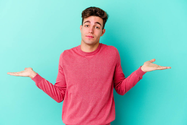 Young caucasian man isolated on blue background doubting and shrugging shoulders in questioning gesture. - Photo, Image
