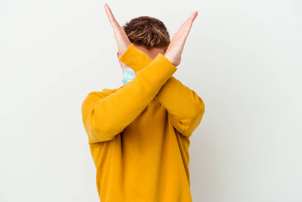 Młody człowiek w masce dla koronawirusa izolowany na białym tle trzymając dwie ręce skrzyżowane, koncepcja zaprzeczenia. - Zdjęcie, obraz