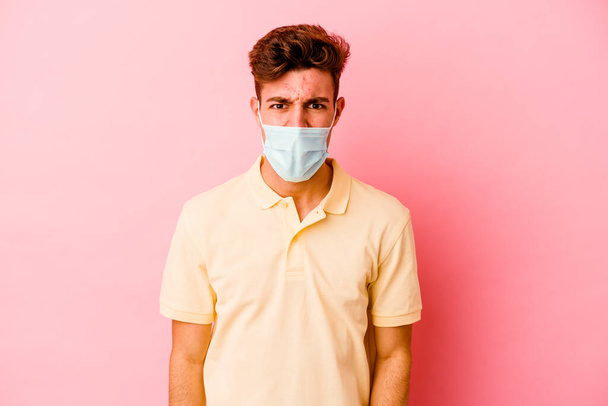 Jovem caucasiano vestindo uma proteção para coronavírus isolado em fundo rosa gritando muito irritado e agressivo. - Foto, Imagem