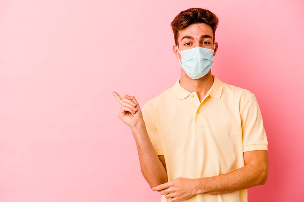 Junger kaukasischer Mann mit einem Coronavirus-Schutz isoliert auf rosa Hintergrund lächelt fröhlich und zeigt mit dem Zeigefinger weg. - Foto, Bild