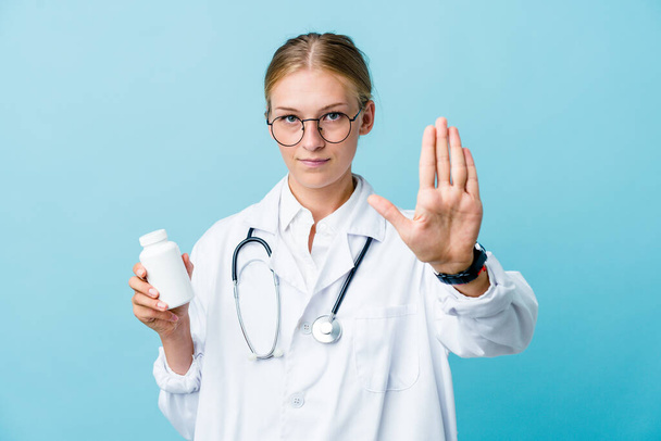 Mladý ruský lékař žena drží pilulky láhev na modrý stojící s nataženou rukou ukazující stop znamení, brání vám. - Fotografie, Obrázek