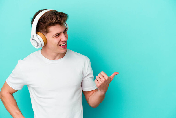 Młody mężczyzna rasy kaukaskiej słuchający muzyki ze słuchawkami odizolowanymi na niebieskim tle palcem kciuka, śmiejący się i beztrosko. - Zdjęcie, obraz