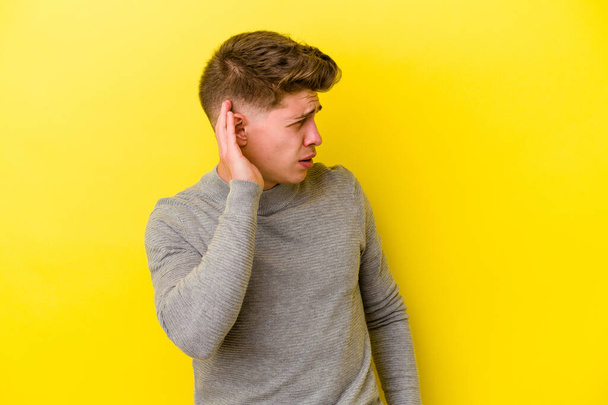 Jeune homme caucasien isolé sur fond jaune essayant d'écouter un potins. - Photo, image