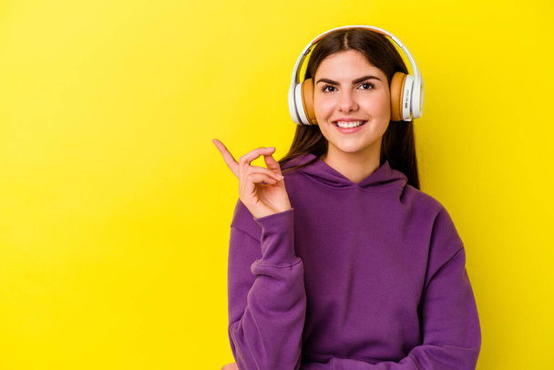 Jonge blanke vrouw luisteren naar muziek met koptelefoon geïsoleerd op roze achtergrond glimlachend vrolijk wijzend met wijsvinger weg. - Foto, afbeelding