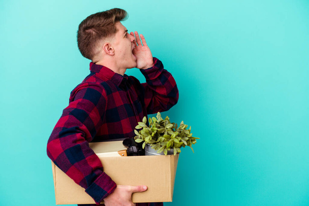 Young caucasian man moving holding a box isolated on blue background shouting and holding palm near opened mouth. - Photo, Image