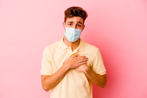 Junger kaukasischer Mann, der einen auf rosa Hintergrund isolierten Coronavirus-Schutz trägt, hat freundlichen Gesichtsausdruck und drückt die Handfläche auf die Brust. Liebeskonzept. - Foto, Bild