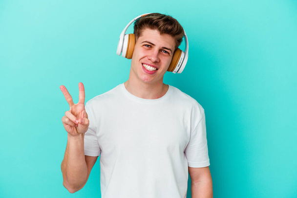Jeune homme caucasien écoutant de la musique avec des écouteurs isolés sur fond bleu joyeux et insouciant montrant un symbole de paix avec les doigts. - Photo, image