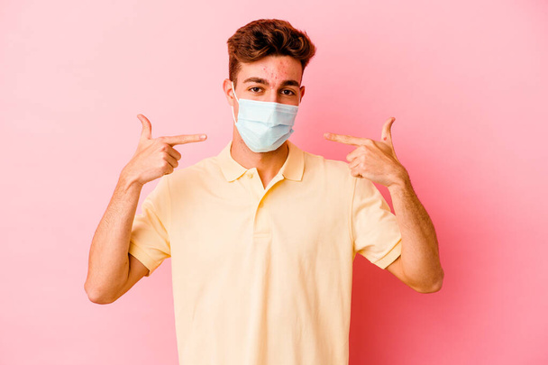 Joven hombre caucásico usando una protección para coronavirus aislado en sonrisas de fondo rosa, señalando con los dedos a la boca. - Foto, Imagen