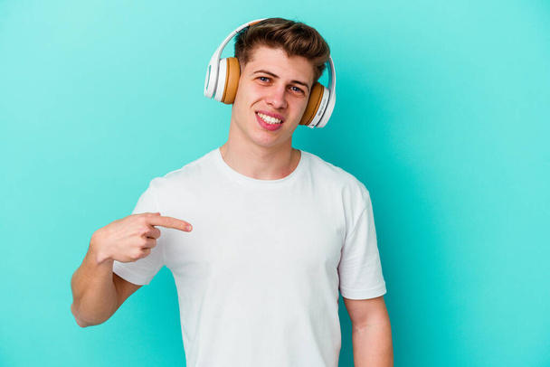 青い背景の人に孤立したヘッドフォンで音楽を聴いている若い白人男性は、シャツのコピースペースを指差して、誇りと自信を持って - 写真・画像