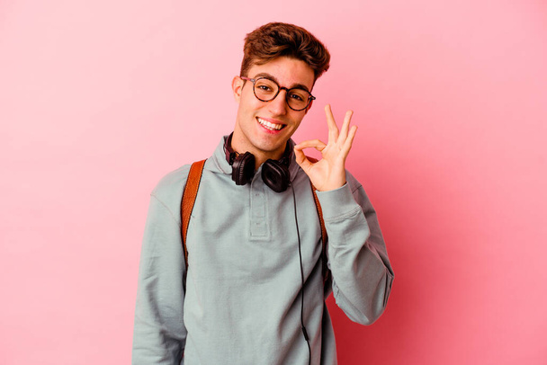 Молодой студент, изолированный на розовом фоне, подмигивает глазом и держит в руках нормальный жест.. - Фото, изображение
