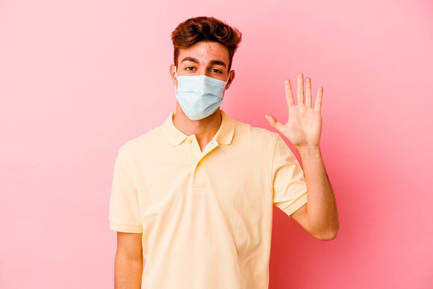 Junger kaukasischer Mann mit einem Coronavirus-Schutz isoliert auf rosa Hintergrund lächelnd fröhlich Nummer fünf mit Fingern zeigend. - Foto, Bild
