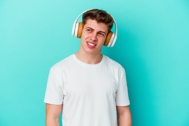 Jonge blanke man die luistert naar muziek met een koptelefoon geïsoleerd op een blauwe achtergrond kijkt opzij glimlachend, vrolijk en aangenaam. - Foto, afbeelding