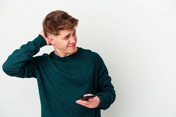 Νεαρός καυκάσιος ξανθός άντρας που χρησιμοποιεί ένα τηλέφωνο απομονωμένο σε λευκό φόντο που αγγίζει το πίσω μέρος του κεφαλιού, σκέφτεται και κάνει μια επιλογή. - Φωτογραφία, εικόνα