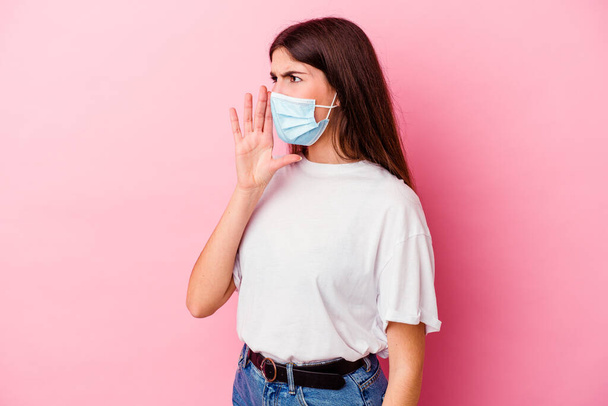 Giovane donna caucasica indossa una maschera per virus isolato su sfondo rosa gridando e tenendo palmo vicino alla bocca aperta. - Foto, immagini