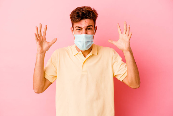 Jovem caucasiano vestindo uma proteção para coronavírus isolado em fundo rosa gritando com raiva. - Foto, Imagem