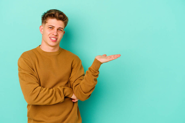 Young caucasian man isolated on blue background showing a copy space on a palm and holding another hand on waist. - Photo, Image