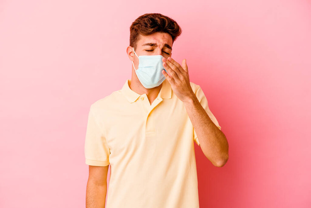 Joven hombre caucásico con protección para coronavirus aislado sobre fondo rosa bostezo mostrando un gesto cansado cubriendo la boca con la mano. - Foto, imagen
