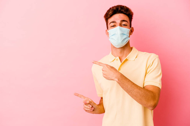Joven hombre caucásico usando una protección para coronavirus aislado sobre fondo rosa excitado señalando con los dedos índice de distancia. - Foto, imagen