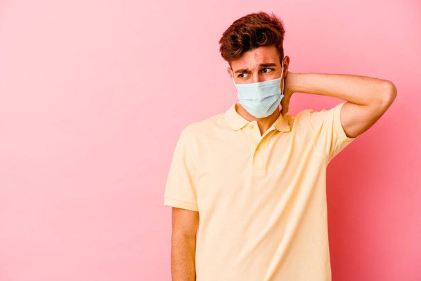 Junger kaukasischer Mann, der einen Coronavirus-Schutz trägt, isoliert auf rosa Hintergrund, berührt den Hinterkopf, denkt nach und trifft eine Entscheidung. - Foto, Bild