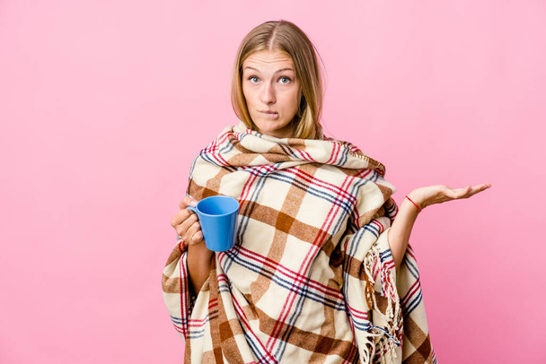 Mladá ruská žena zabalená v pokrývce pití kávy zmatené a pochybné pokrčení ramen držet kopii prostor. - Fotografie, Obrázek