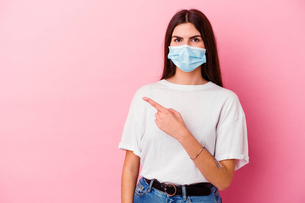 Young caucasian woman wearing a mask for virus isolated on pink background smiling and pointing aside, showing something at blank space. - Foto, imagen