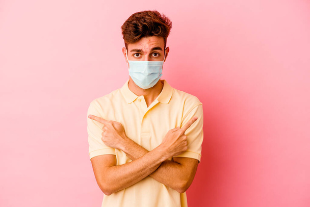 Junge kaukasische Mann trägt einen Schutz für Coronavirus isoliert auf rosa Hintergrund Punkte seitlich, versucht, zwischen zwei Optionen zu wählen. - Foto, Bild