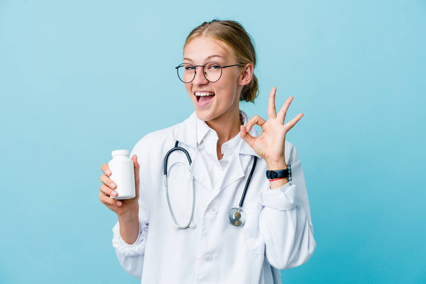 Young russian doctor woman holding pills bottle on blue winks an eye and holds an okay gesture with hand. - Photo, Image