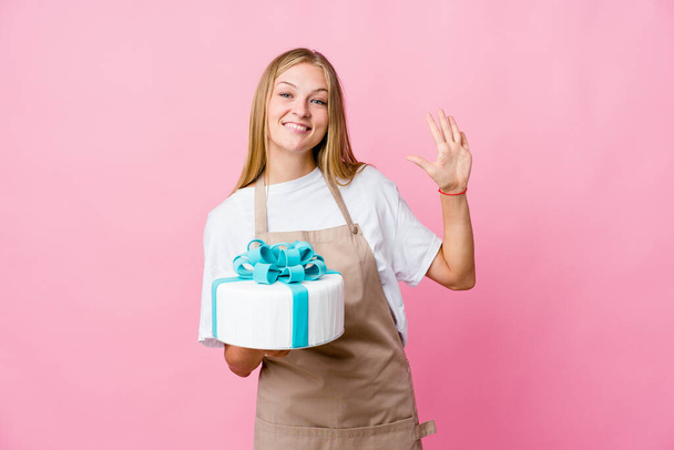 Νεαρή Ρωσίδα φούρναρης κρατώντας ένα νόστιμο κέικ χαμογελώντας χαρούμενα δείχνει το νούμερο πέντε με τα δάχτυλα. - Φωτογραφία, εικόνα
