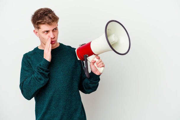 Young caucasian man holding a megaphone isolated on white background is saying a secret hot braking news and looking aside - Foto, imagen
