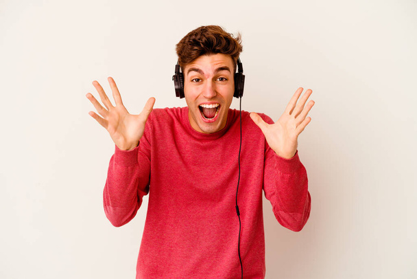 Young caucasian man listening to music isolated on white background celebrating a victory or success, he is surprised and shocked. - Photo, Image