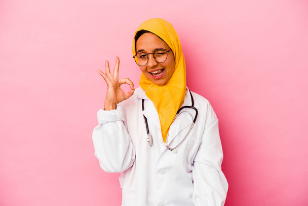 Junge muslimische Ärztin, isoliert auf rosa Hintergrund, zwinkert mit einem Auge und hält eine Okay-Geste mit der Hand. - Foto, Bild