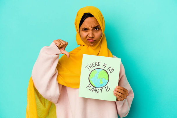 Junge muslimische Frau mit einem Plakat auf blauem Hintergrund, das eine ablehnende Geste zeigt, Daumen nach unten. Konzept der Uneinigkeit. - Foto, Bild