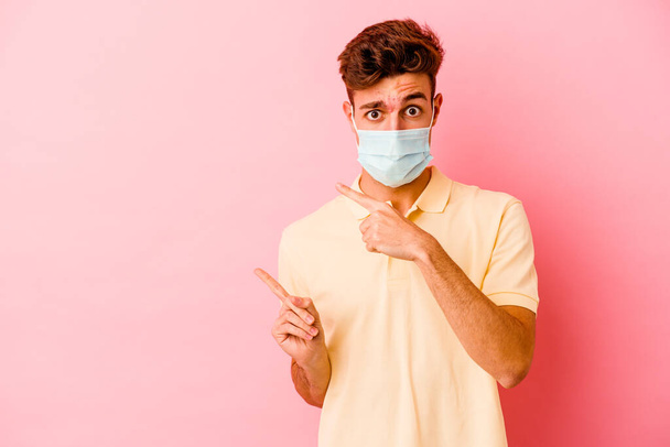 Junger kaukasischer Mann mit Coronavirus-Schutz isoliert auf rosa Hintergrund schockiert mit Zeigefinger auf Kopierraum zeigend. - Foto, Bild