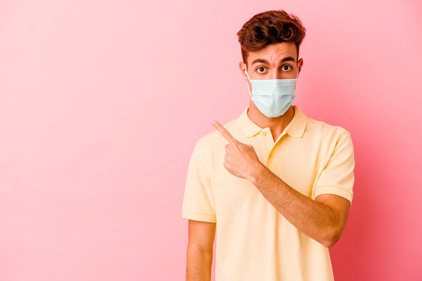 Jonge blanke man draagt een bescherming voor coronavirus geïsoleerd op roze achtergrond wijzend naar de zijkant - Foto, afbeelding